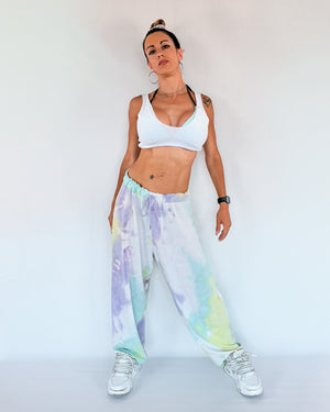Woman wearing a white sports bra and tie-dye oversized dance pants on a plain background